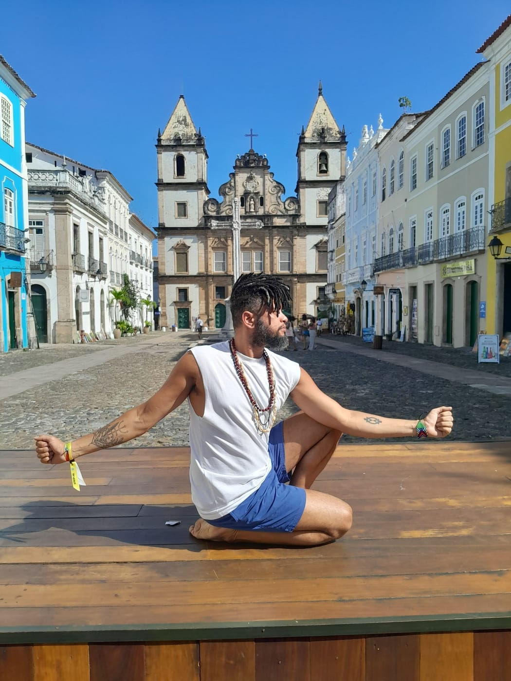 Keâ Costa em postura sentada, com os braços abertos, em uma praça histórica